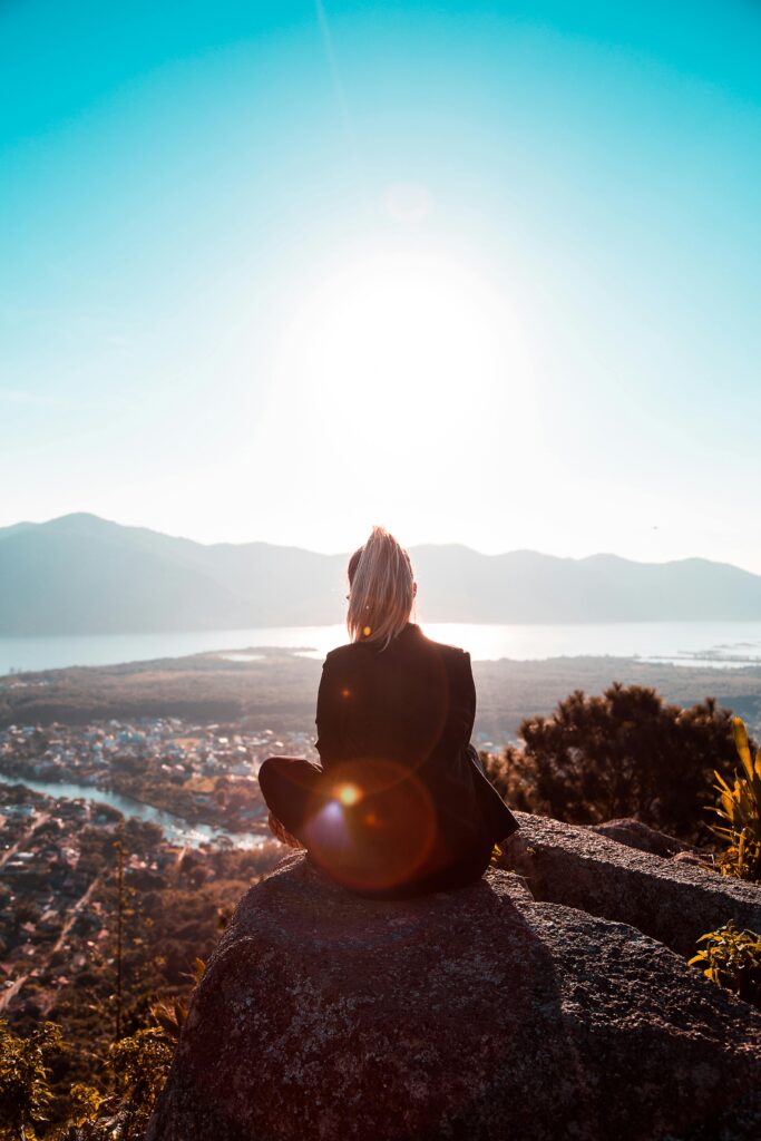 A woman sits on a cliff, gazing at a stunning valley landscape under a bright sun.